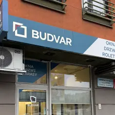 Logo firmy BUDVAR z napisem "OKNA DRZWI ROLETY" na tle pomarańczowej ściany budynku.